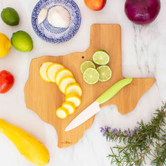 A Totally Bamboo Destination Texas Cutting Board holds sliced yellow squash and lime halves, with a knife featuring a green handle resting beside them. Surrounding the board on the marble surface are limes, a lemon, a tomato, garlic on a small plate, and a red onion—a perfect touch of Texas kitchen decor.