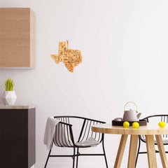 Modern kitchen showcasing Texas kitchen decor, featuring a round wooden table with black chairs and a white wall adorned with the Totally Bamboo Destination Texas Cutting Board, 14" x 13-3/8", serving as an eye-catching wooden art piece. A teapot and two lemons rest on the table. A light wood cabinet and a geometric vase filled with greenery complete this stylish setting.