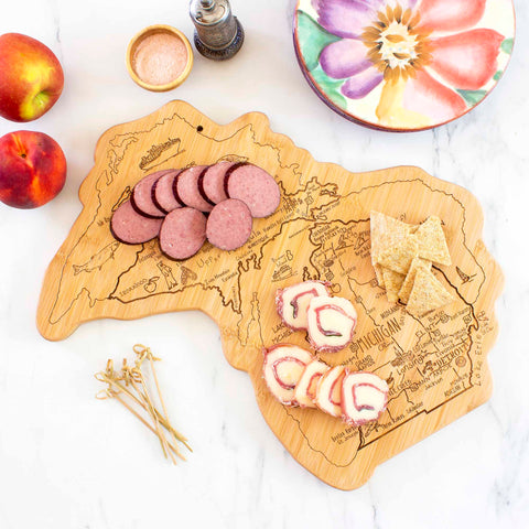 The Destination Michigan Cutting Board by Totally Bamboo offers a delightful spread featuring sliced sausage, crackers, and rolled ham with cheese. Beside this state-shaped bamboo cutting surface, two apples rest alongside a small bowl of salt, a pepper grinder, and a colorful flower-patterned plate on marble.