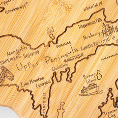 The Totally Bamboo "Destination Michigan Cutting Board" intricately features the Upper Peninsula of Michigan with engravings of locations such as Marquette, Iron Mountain, and Munising, while a ship illustration embellishes Beaver Island. This cutting board is a part of our Destination Series and is expertly crafted on a light wood surface, measuring 16-3/4" x 16-1/2".