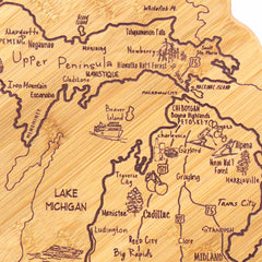 The Totally Bamboo Destination Michigan Cutting Board, measuring 16-3/4" x 16-1/2", is intricately designed to represent the state of Michigan and features both the Upper and Lower Peninsulas. It artistically includes hand-drawn depictions of towns, roads, and landmarks, prominently showcasing Lake Michigan on one side along with Mackinac Island, Petoskey, Traverse City, and Cadillac.