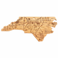 Totally Bamboo's Destination North Carolina Cutting Board, measuring 18" x 8", is a state-shaped wooden board showcasing laser-engraved designs with city names and local landmarks such as Asheville, Raleigh, and Charlotte. Decorative illustrations highlight regional attractions and notable sites throughout the state.