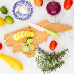 The Destination North Carolina Cutting Board by Totally Bamboo, with its striking bamboo cutting surface, elegantly showcases sliced lemons and limes. It's accompanied by an assortment of fresh vegetables, including onion, tomatoes, squash, limes, and garlic with rosemary sprigs and a small knife. A blue decorative plate is placed nearby on the marble surface.