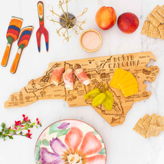 The Destination North Carolina Cutting Board by Totally Bamboo, measuring 18" x 8", artistically showcases prosciutto, cheese slices, and pickles. Encircling this unique bamboo cutting surface are crackers, peaches, colorful spoons and a fork, a small bowl of salt, and a floral plate on a marble surface.