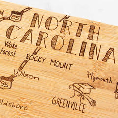 The Totally Bamboo Destination North Carolina Cutting Board, measuring 18" x 8", is exquisitely designed with a laser-engraved map of the state. It highlights cities such as Rocky Mount, Greenville, Wilson, Wake Forest, and Plymouth and includes charming decorative road signs and markings.