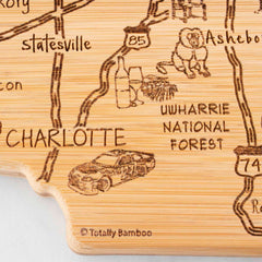 The "Destination North Carolina" cutting board by Totally Bamboo measures 18" x 8" and is crafted from wood in the shape of the state. It features laser-engraved images of race cars, a basket of apples, a lion, and roads across North Carolina, including Charlotte, Statesville, and Uwharrie National Forest. The bottom includes the brand's signature engraving.