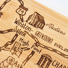 Close-up of a laser-engraved artwork on the Totally Bamboo Destination North Carolina Cutting Board, 18" x 8". The board is shaped like the state and highlights cities such as Winston-Salem, Greensboro, and High Point with illustrated landmarks and roads including Route 74.