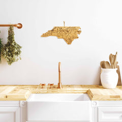 A kitchen boasting a wooden counter, white sink, and copper faucet showcases a bamboo cutting surface and spoon holder. Hanging above is the Destination North Carolina Cutting Board by Totally Bamboo, measuring 18" x 8", perfectly complemented by herbs suspended on the left side.