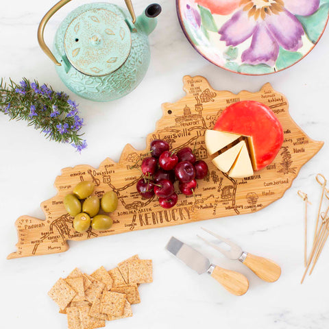 The Destination Kentucky Cutting Board by Totally Bamboo provides a charming bamboo surface for slicing cheese, cherries, and olives. Surrounding it are cheese knives, crackers, a teal teapot, a floral ceramic bowl, and sprigs of rosemary elegantly arranged on a white marble surface.