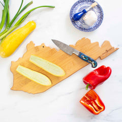 A Destination Kentucky Cutting Board by Totally Bamboo, measuring 18" x 8-1/2", holds cut yellow squash, while a knife lies nearby. A halved red bell pepper rests on the table. Completing the scene, a blue and white plate showcases a garlic bulb, a garlic press, and green onions in harmonious culinary wall art.