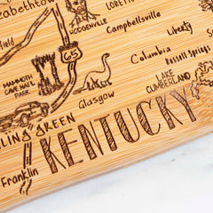 Totally Bamboo presents the Destination Kentucky Cutting Board, measuring 18" x 8-1/2", crafted from wood to serve as distinctive wall art. It features an engraved map of Kentucky with cities including Bowling Green, Glasgow, and Franklin, along with illustrations like a dinosaur for Mammoth Cave National Park and other local landmarks.