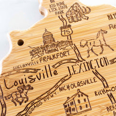 The Destination Kentucky Cutting Board by Totally Bamboo, measuring 18" x 8-1/2", makes a perfect piece of wall art with its engraved illustrations featuring landmarks like the state capitol, a horse, and historical sites. It prominently marks city names such as Lexington and Louisville.