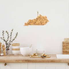 A kitchen counter showcases an 18" x 8-1/2" Destination Kentucky Cutting Board by Totally Bamboo, along with bowls and salt and pepper grinders. A decorative wooden map of Kentucky graces the white wall as art, beautifully complemented by a small branch in a vase for added decor.