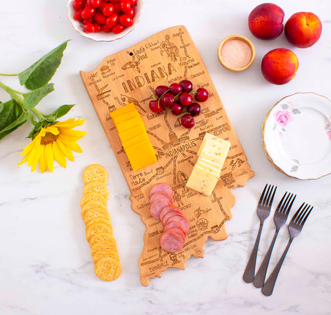 The Totally Bamboo Destination Indiana Cutting Board, measuring 15-3/4" x 10", features a beautifully laser-engraved outline of Indiana and serves as an elegant display for cheese, cherries, and sliced meat. It is elegantly arranged alongside crackers, tomatoes, peaches, a sunflower, a floral plate filled with salt, and four forks on an immaculate white surface.