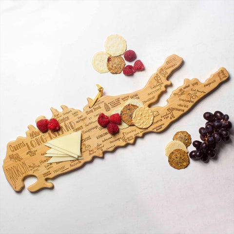 The Totally Bamboo Destination Long Island Cutting Board, measuring 22" x 6-1/2", features laser-engraved city names and is shaped like Long Island. It elegantly holds cheese slices, crackers, and raspberries, while resting on a bamboo cutting surface that is adorned with additional crackers, raspberries, and a bunch of grapes against a pristine white background.