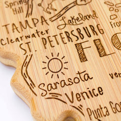 A close-up of the Destination Florida Cutting Board by Totally Bamboo reveals a state-shaped decorative wooden design engraved with the names of several Florida cities, such as Tampa, Clearwater, St. Petersburg, Sarasota, Venice, and Lakeland. The charming piece from the Destination Series also features illustrations like a sun and palm trees alongside the text.