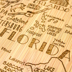 A detailed view of the Destination Florida Cutting Board by Totally Bamboo showcases laser-engraved cities and landmarks such as Orlando, Kissimmee, and Lake Okeechobee. This intricate piece, measuring 13-1/2" x 12-1/4", adds a rustic and artistic flair to any decor.