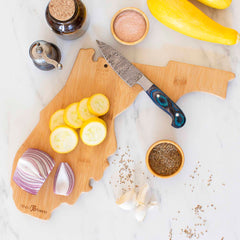 On a marble surface, the Destination Florida Cutting Board by Totally Bamboo is artfully presented with a knife and freshly sliced yellow squash and red onion. The uniquely shaped wooden board also features intricate laser-engraved artwork of Florida, while nearby are whole garlic cloves, small bowls of spices, and whole yellow squash.