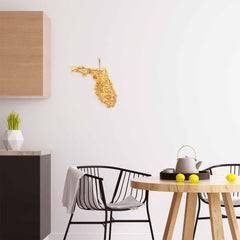 A modern dining area includes a black chair, a light wood table, and a teapot with lemons. The white wall features the Totally Bamboo Destination Florida Cutting Board, measuring 13-1/2" x 12-1/4", hanging next to a kitchen cabinet. A gray towel drapes over the chair while a small plant sits on the counter.