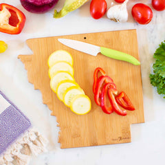 Discover the Totally Bamboo Destination Arkansas Cutting Board, measuring 13" x 12". This beautifully crafted wooden board is shaped like the state of Arkansas and features a display of sliced yellow squash and red bell pepper. Accompanying the setup is a white knife with a green handle, while tomatoes, garlic, lettuce, corn, and a purple-patterned cloth elegantly encircle this bamboo cutting surface.