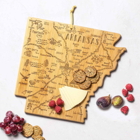 The Destination Arkansas Cutting Board by Totally Bamboo, measuring 13" x 12", is a bamboo-crafted item shaped like the state of Arkansas and features engravings of city names and landmarks. On a white countertop, it beautifully presents cheese, crackers, figs, grapes, and raspberries.