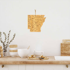 A cozy kitchen scene features the Totally Bamboo Destination Arkansas Cutting Board, measuring 13" x 12", hanging on a white wall. Below, a countertop holds ceramic bowls, a wooden spoon, and a bamboo cutting surface adorned with a sprig of greenery in a vase.