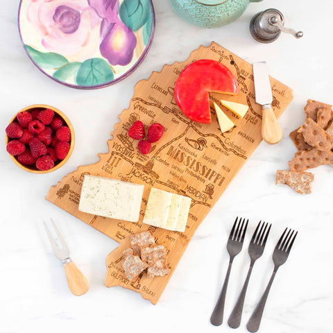 A Totally Bamboo Destination Mississippi Cutting Board is elegantly arranged on a marble surface, featuring an assortment of cheeses, crackers, and raspberries. Nearby, there is a bowl of raspberries accompanied by a knife, a cheese fork, and four forks. In the background, you can see a decorative flower plate and a patterned bowl with intricate laser-engraved artwork.