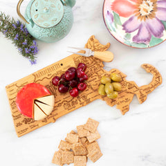 A Totally Bamboo Destination Massachusetts Cutting Board, measuring 16-3/4" x 9-1/4", holds an assortment of cheese, cherries, and olives on its bamboo surface. A cheese knife is elegantly positioned on the board. In close proximity are crackers, a vibrant teapot in green hues, and a colorful plate. Adding a touch of charm to the setting are sprigs of lavender placed next to the teapot.