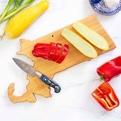 A Totally Bamboo Destination Massachusetts Cutting Board, measuring 16-3/4" x 9-1/4", holds sliced red pepper and halved yellow squash. A decorative knife rests beside the vegetables, while spring onions and a whole red pepper sit on the marble countertop. This state-shaped board features laser-engraved artwork, adding charm to your kitchen.