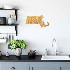 In a modern kitchen, a sleek black countertop and gray cabinets are beautifully contrasted with a white sink. A potted plant adds vitality near the bamboo cutting surface. Above, various kitchen items rest alongside the Totally Bamboo Destination Massachusetts Cutting Board (16-3/4" x 9-1/4"), which features laser-engraved artwork of Massachusetts in the shape of the state, bringing local charm to the space.