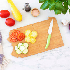 Totally Bamboo's Destination Pennsylvania Cutting Board, measuring 16" x 9-3/4", features a beautifully arranged display of sliced tomatoes, yellow squash, and zucchini on its bamboo surface. Accompanied by a knife with a green handle from the Destination Series, this culinary scene is set alongside whole tomatoes, a garlic bulb, fresh basil leaves, salt, and a pepper grinder.