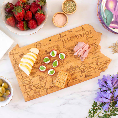 The Destination Pennsylvania Cutting Board by Totally Bamboo, measuring 16" x 9-3/4", elegantly displays cheese, crackers, green-wrapped candies, and ham slices. Positioned on a marble surface, it is artistically surrounded by strawberries, olives, and a vase of purple flowers on this laser-engraved bamboo cutting board.