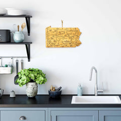 A modern kitchen features light blue cabinets, a white countertop, and a silver faucet. Accenting the space are a potted plant and decorative bowl alongside a bamboo cutting surface by Totally Bamboo. Hanging above is the Destination Pennsylvania Cutting Board, 16" x 9-3/4", showcasing exquisite laser-engraved artwork from their series.