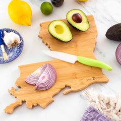 On a Totally Bamboo Destination Louisiana Cutting Board, measuring 13" x 12", sliced avocado and red onion are artfully arranged. A ceramic knife with a green handle lies on the board, while nearby lemons, garlic, lime, a brush, and a decorative plate are placed on the marble countertop.