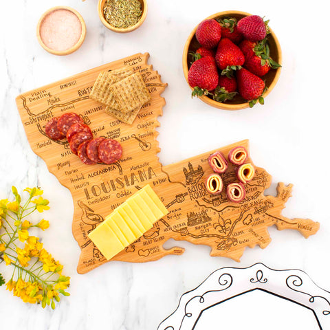 The Totally Bamboo Destination Louisiana Cutting Board, measuring 13" x 12", gracefully presents cheese slices, crackers, and salami. Dishes filled with strawberries and pinwheel sandwiches are placed nearby. Yellow flowers are tastefully arranged to the side on a spotless white tabletop.