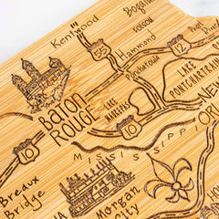 The Destination Louisiana Cutting Board by Totally Bamboo is a bamboo state-shaped board, elegantly laser-engraved with a stylized map of Louisiana that highlights Baton Rouge, New Orleans, major highways, landmarks such as Lake Pontchartrain, and features a fleur-de-lis design. Nearby routes and cities are clearly labeled for added detail. The cutting board measures 13" x 12".