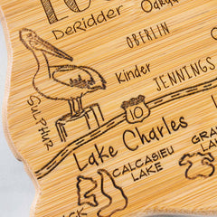 A detailed view of the "Destination Louisiana Cutting Board" by Totally Bamboo, measuring 13" x 12", features a laser-engraved pelican perched on a post. The design intricately highlights Lake Charles and neighboring areas such as Sulphur, Kinder, and Jennings. It also includes the Interstate 10 sign and Calcasieu Lake etched into the bamboo surface.