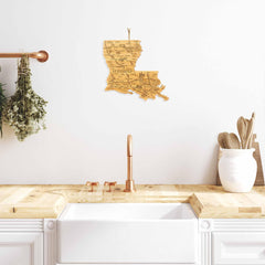 A modern kitchen featuring a white farmhouse sink with a wooden countertop and a Totally Bamboo Destination Louisiana Cutting Board, measuring 13" x 12". A map of Louisiana hangs on the white wall above, complemented by laser-engraved artwork. Copper fixtures and potted herbs accentuate the space, alongside nearby wooden utensils.