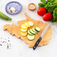 A Destination West Virginia Cutting Board from Totally Bamboo, measuring 13-1/2" x 11-3/4", features a design inspired by the state of West Virginia and holds sliced yellow and green zucchini. Beside it are a knife, lettuce, tomatoes, zucchini, peppercorns, salt in a small bowl, and a decorative blue and white plate adorned with garlic.