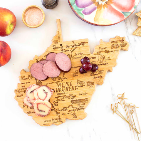 Displayed on the marble surface is the Destination West Virginia Cutting Board by Totally Bamboo, shaped distinctively in state form and crafted from bamboo. It features laser-engraved city names and currently holds slices of sausage, pimento cheese, and small red fruits. Beside it are apples, a bowl of salt, breadsticks, and decorative skewers.