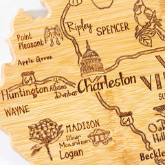 The Totally Bamboo Destination West Virginia Cutting Board, measuring 13-1/2" x 11-3/4", features laser-engraved artwork of Charleston, Huntington, and Spencer. It includes intricate symbols such as the Capitol building, trees, and roads marked G64, effectively melding art with functionality similar to state-shaped cutting boards.