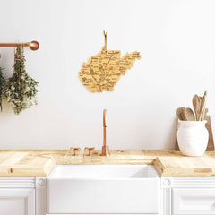 A wooden map of West Virginia is displayed above the kitchen sink on a white wall. The countertop features the "Destination West Virginia Cutting Board" by Totally Bamboo, alongside other state-shaped cutting boards and utensils, with a copper faucet perfectly centered. Hanging dried herbs are complemented by laser-engraved artwork, enhancing the charm of this cozy space.