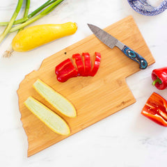 The Totally Bamboo Destination Nebraska Cutting Board, measuring 17-3/4" x 9", showcases laser-engraved artwork and holds sliced red bell peppers and yellow squash. A knife with a blue-green handle is placed on the board, while scallions and additional peppers adorn the marble countertop nearby.