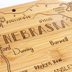 The Destination Nebraska Cutting Board by Totally Bamboo offers a bamboo cutting surface adorned with laser-engraved artwork depicting the map of Nebraska, highlighting towns like Ainsworth and O'Neill, as well as notable landmarks such as Ashfall Fossil Beds.