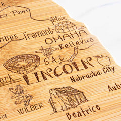 The Destination Nebraska Cutting Board by Totally Bamboo, measuring 17-3/4" x 9", is a state-shaped cutting board made from bamboo. It features an engraved map of Nebraska cities such as Omaha and Lincoln, with laser-engraved artwork highlighting landmarks like a stadium and a log cabin, all enhanced with ornate lettering and decorative elements.