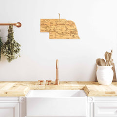 A sleek kitchen features a white sink, wooden countertop, and copper faucet. Above the sink on the white wall is a laser-engraved Destination Nebraska Cutting Board by Totally Bamboo, measuring 17-3/4" x 9". Dried herbs adorn the left side, while utensils and a bamboo cutting surface are neatly arranged to the right.