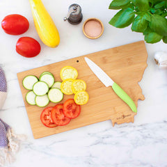 A Destination Montana Cutting Board from Totally Bamboo, measuring 16" x 9-1/2" and shaped like the state of Montana, holds sliced tomatoes, zucchini, and yellow squash. A green-handled knife is placed nearby. The marble surface is also adorned with whole tomatoes, a squash, salt, a pepper shaker, garlic cloves, and basil leaves.