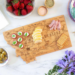 A Totally Bamboo Destination Montana Cutting Board, measuring 16" x 9-1/2", beautifully presents ham slices, apple wedges, round crackers, and mini caprese bites. Surrounding the board are bowls filled with strawberries, olives, other snacks, and purple flowers. The laser-engraved artwork adds a unique touch to your spread.