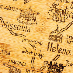 The Totally Bamboo Destination Montana Cutting Board, measuring 16" x 9-1/2", features a laser-engraved wooden map with labeled locations, such as Missoula, Helena, Anaconda, Drummond, Deer Lodge, and Butte. It includes rustic-style illustrations of a carousel, scenic landscapes, and historic buildings.