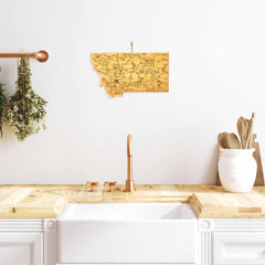 A Destination Montana Cutting Board by Totally Bamboo, measuring 16" x 9-1/2", hangs on a white wall above a farmhouse sink with a copper faucet. To the left, bundles of herbs are hung to dry. On the right, a bamboo cutting board and wooden container hold utensils. The countertop is made of light wood.